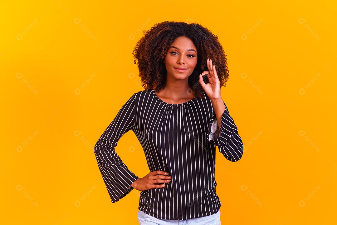 Linda garota de cabelos cacheados usando as mãos okey sinal. Fundo amarelo. mulher afro ok