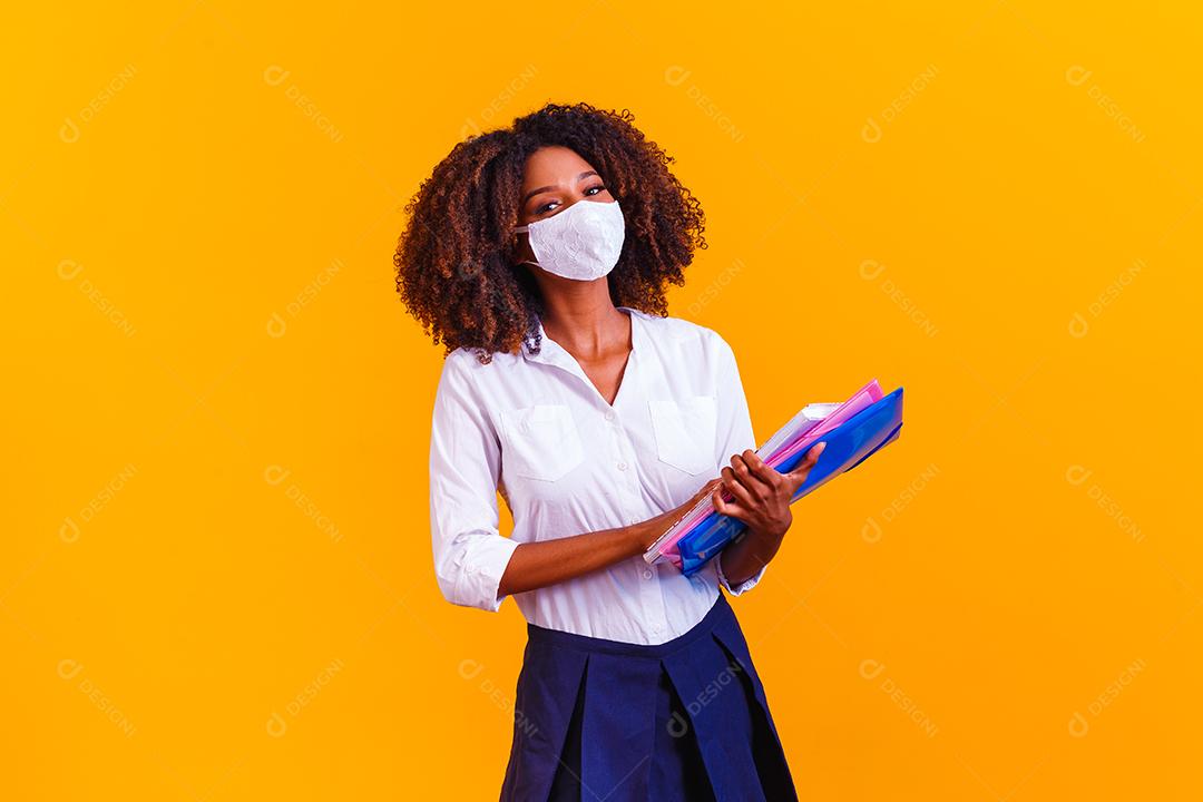 Garota afro segurando livros em fundo amarelo usando máscara protetora. Retorno às aulas por quarentena