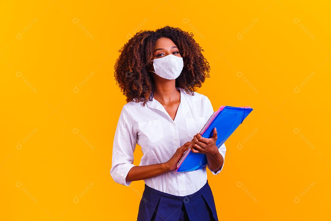 Garota afro segurando livros em fundo amarelo usando máscara protetora. Retorno às aulas por quarentena