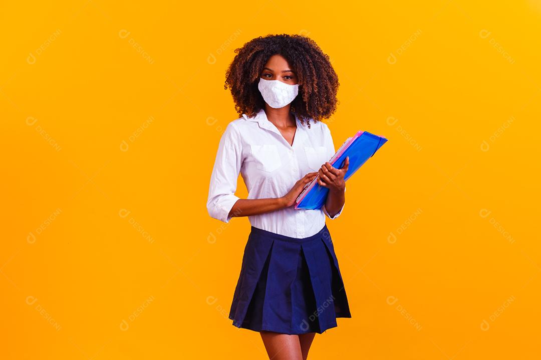 Garota afro segurando livros em fundo amarelo usando máscara protetora. Retorno às aulas por quarentena