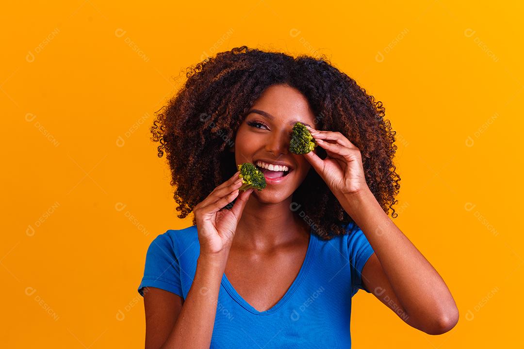 mulher afro feliz comendo brócolis. mulher com brócolis em fundo amarelo. conceito de alimentação saudável