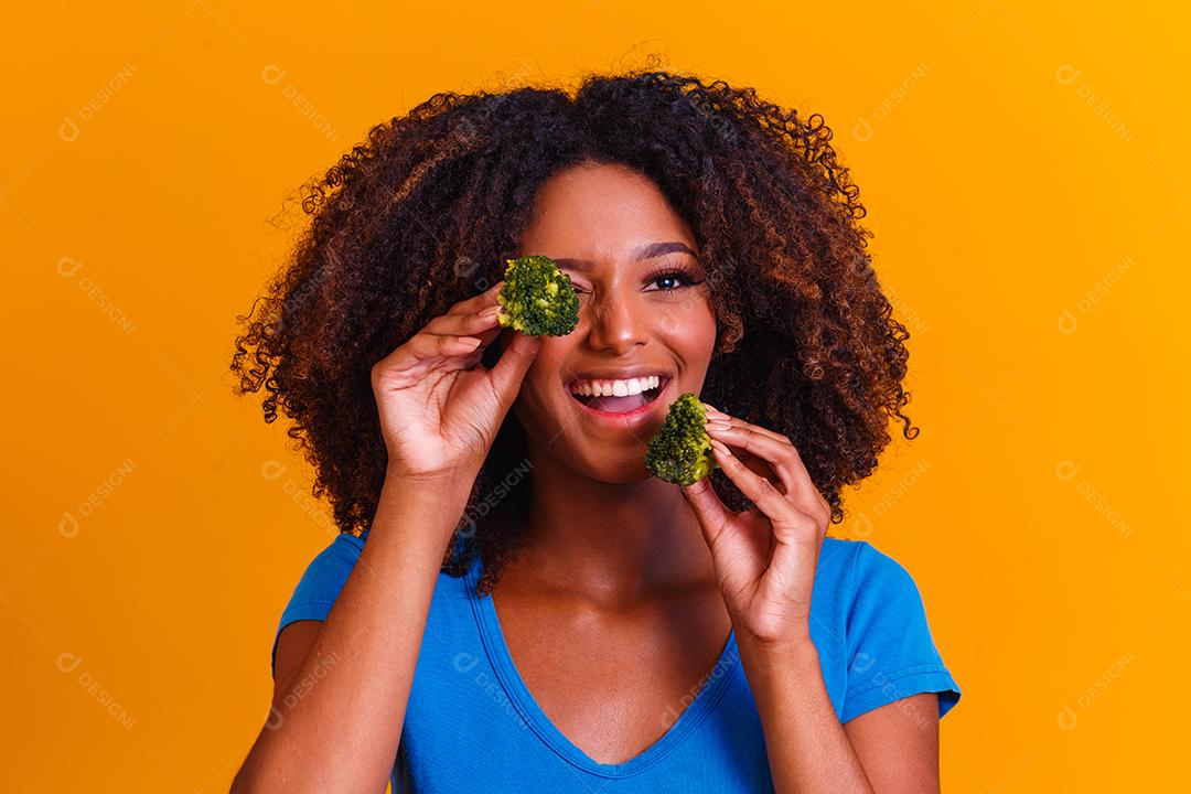 mulher afro feliz comendo brócolis. mulher com brócolis em fundo amarelo. conceito de alimentação saudável