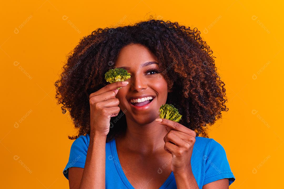 mulher afro feliz comendo brócolis. mulher com brócolis em fundo amarelo. conceito de alimentação saudável