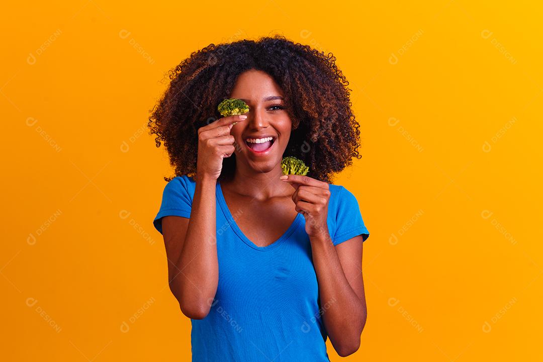 mulher afro feliz comendo brócolis. mulher com brócolis em fundo amarelo. conceito de alimentação saudável