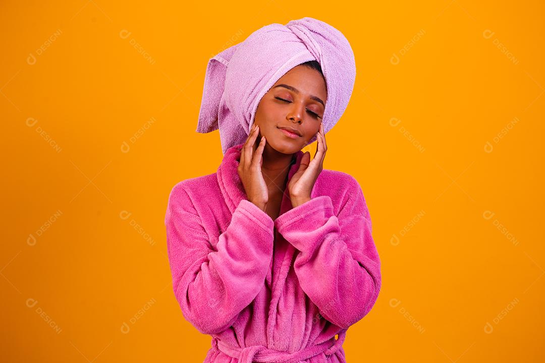 Mulher afro com toalha na cabeça e roupão de banho depois de sair do banho em fundo amarelo. Conceito de banho e cuidados