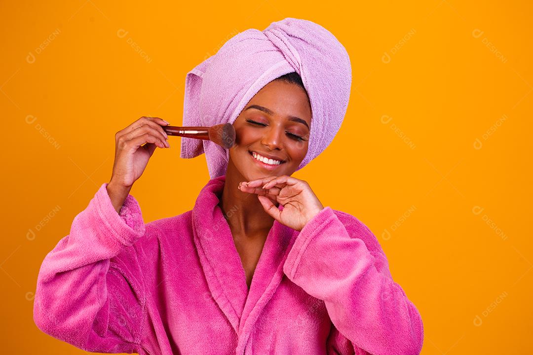 Mulher afro com toalha na cabeça e roupão de banho depois de sair do banho em fundo amarelo. Conceito de banho e cuidados