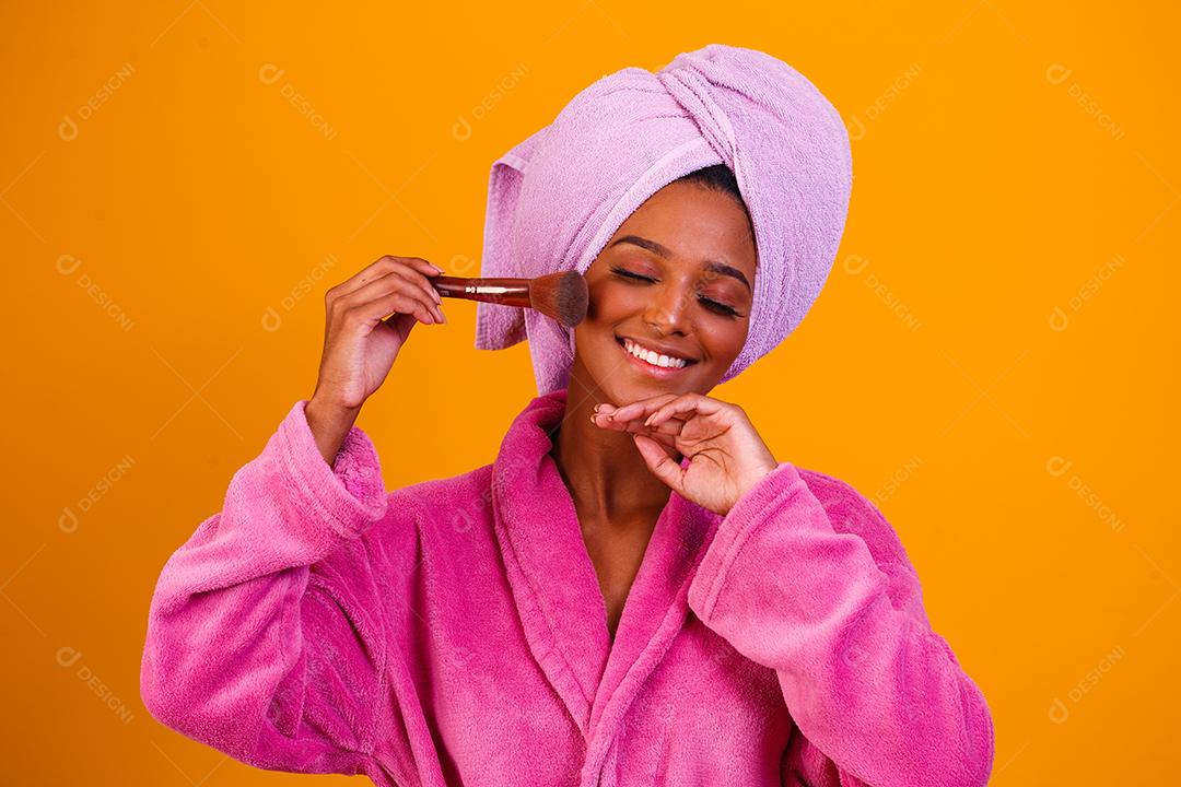 Mulher afro com toalha na cabeça e roupão de banho depois de sair do banho em fundo amarelo. Conceito de banho e cuidados
