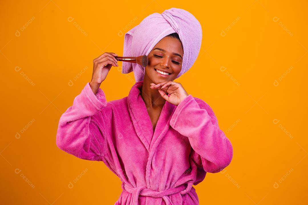Mulher afro com toalha na cabeça e roupão de banho depois de sair do banho em fundo amarelo. Conceito de banho e cuidados