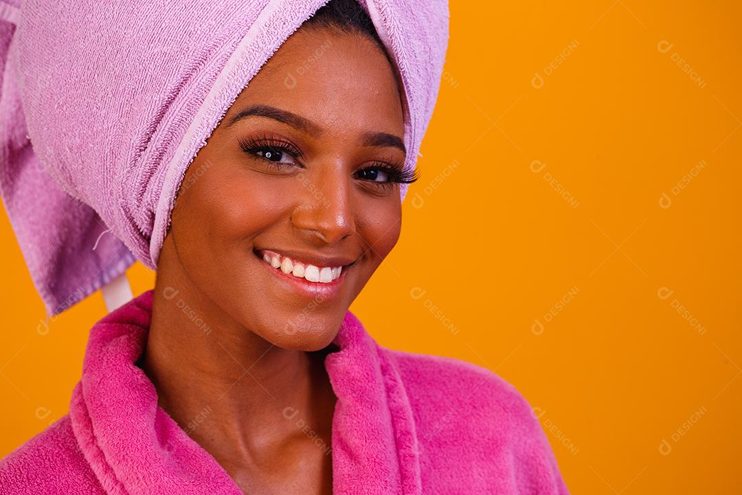 Mulher afro com toalha na cabeça e roupão de banho depois de sair do banho em fundo amarelo. Conceito de banho e cuidados