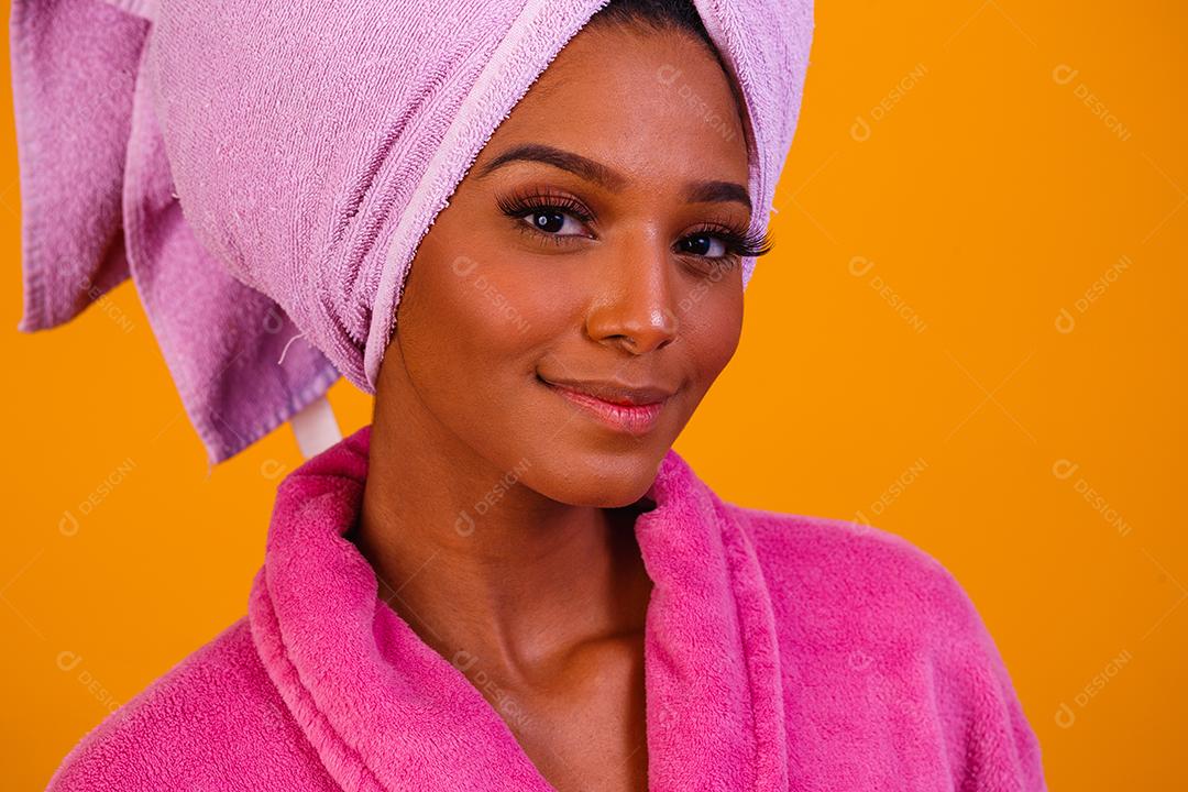 Mulher afro com toalha na cabeça e roupão de banho depois de sair do banho em fundo amarelo. Conceito de banho e cuidados