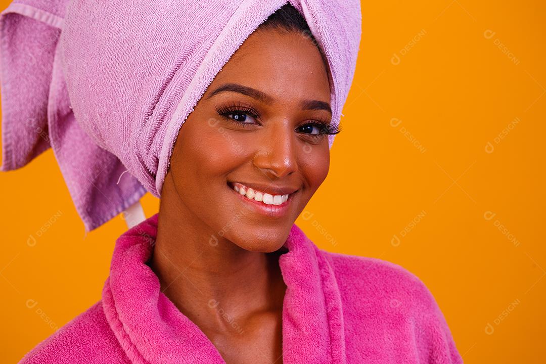Mulher afro com toalha na cabeça e roupão de banho depois de sair do banho em fundo amarelo. Conceito de banho e cuidados