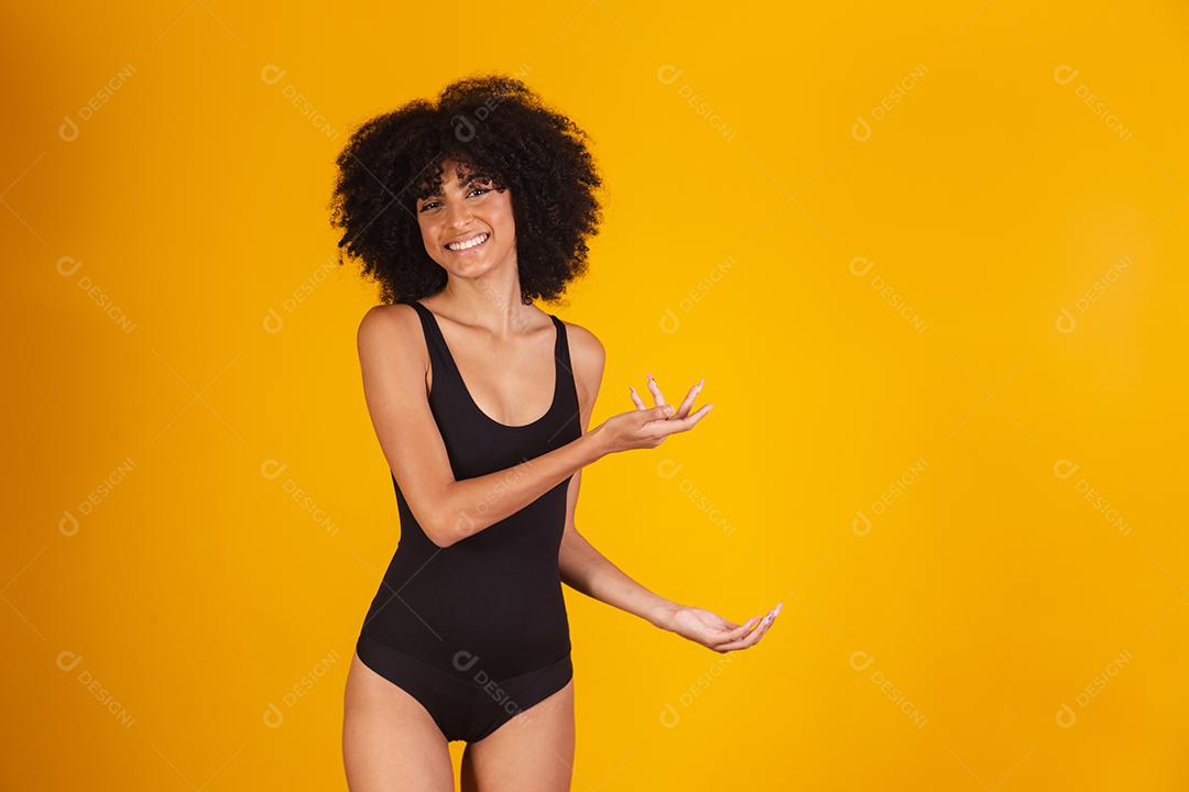 Mulher bonita com cabelo afro em traje de banho em fundo amarelo, apontando para o lado