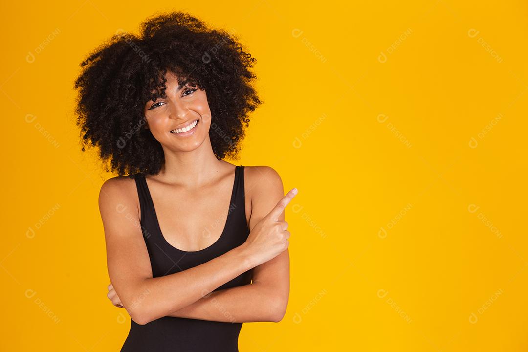 Mulher bonita com cabelo afro em traje de banho em fundo amarelo, apontando para o lado