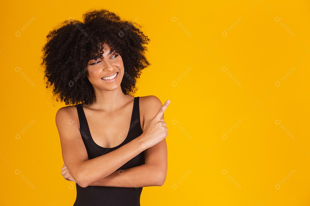 Mulher bonita com cabelo afro em traje de banho em fundo amarelo, apontando para o lado