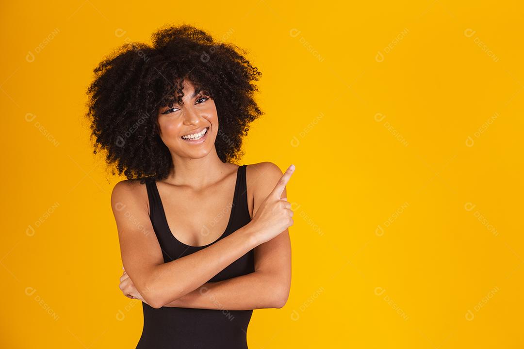Mulher bonita com cabelo afro em traje de banho em fundo amarelo, apontando para o lado