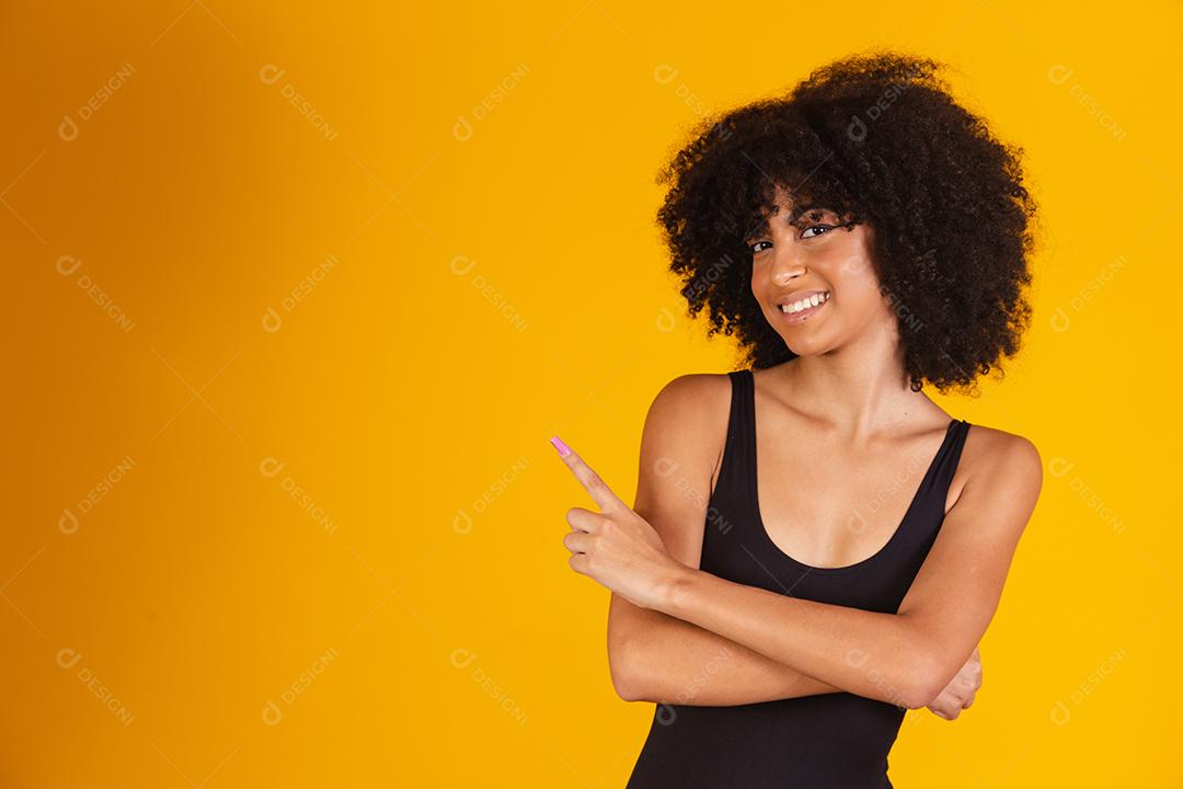 Mulher bonita com cabelo afro em traje de banho em fundo amarelo, apontando para o lado