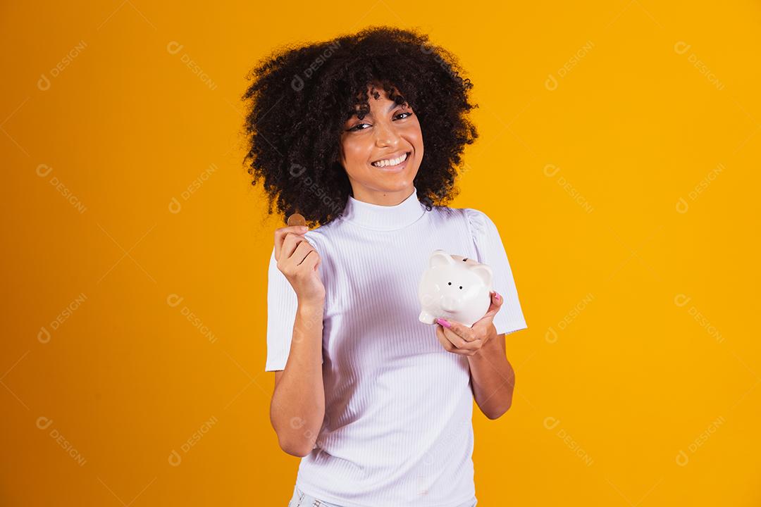 Mulher negra segurando um cofrinho. Mulher economizando dinheiro em porquinho.