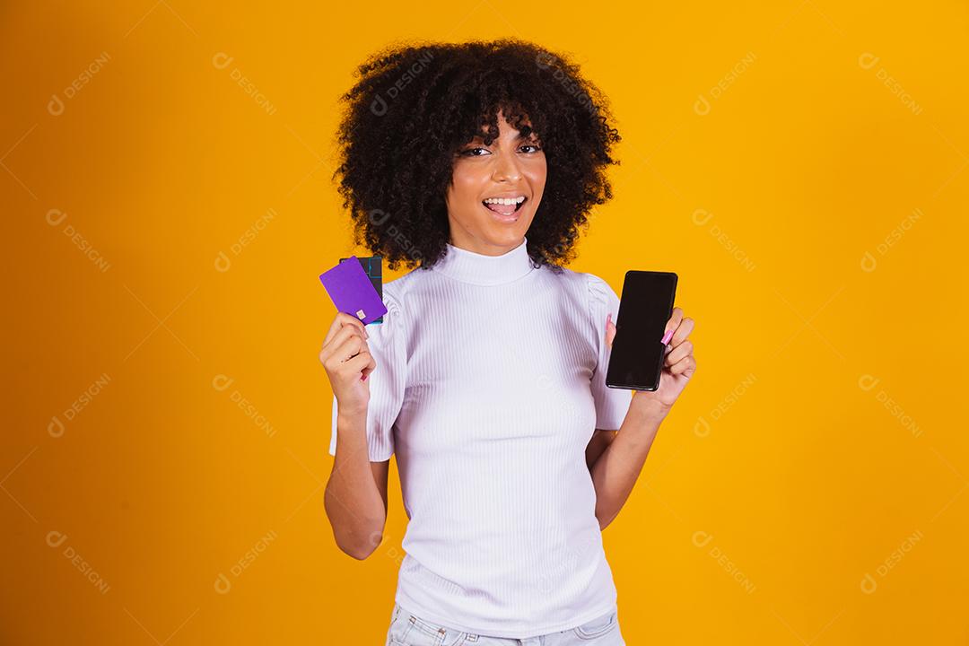 Foto de linda mulher de pele escura animada segurando cartão de crédito plástico mostrando novo modelo de telefone