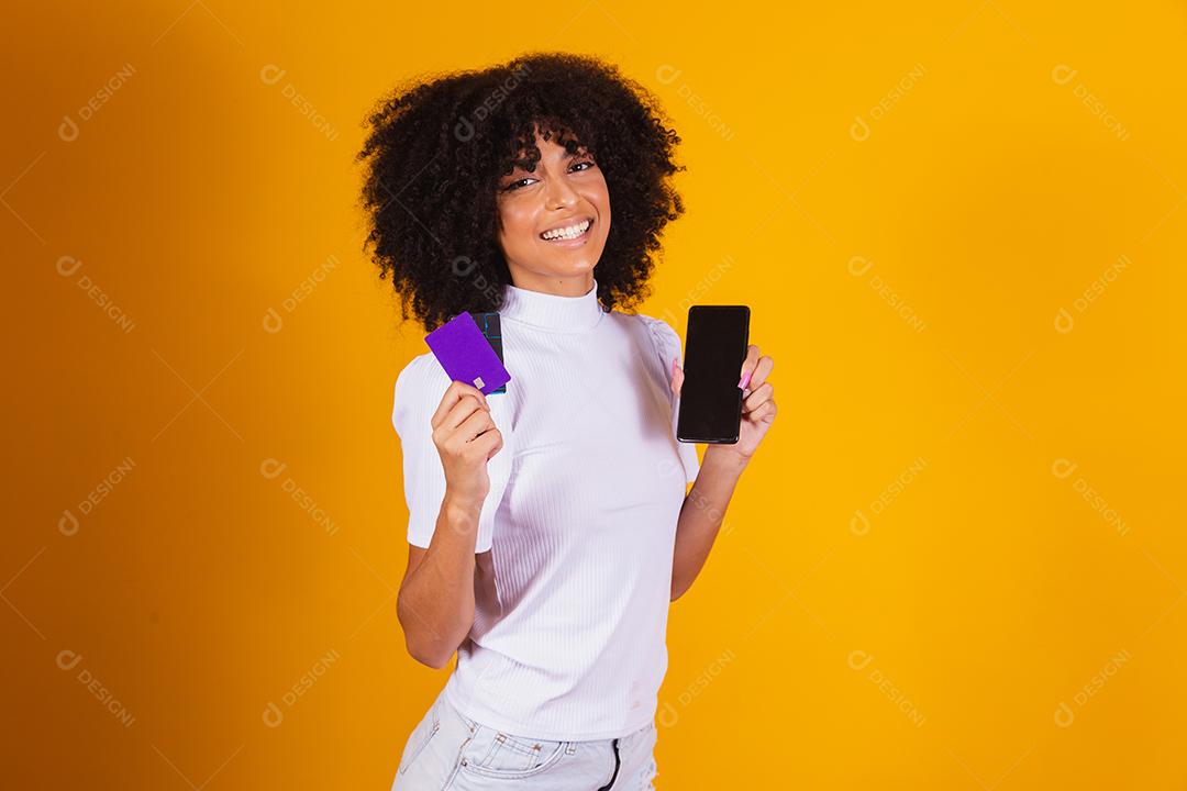 Foto de linda mulher de pele escura animada segurando cartão de crédito plástico mostrando novo modelo de telefone