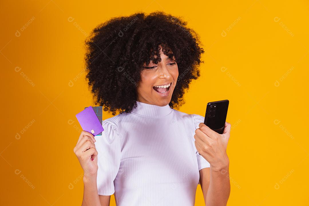 Foto de linda mulher de pele escura animada segurando cartão de crédito plástico mostrando novo modelo de telefone