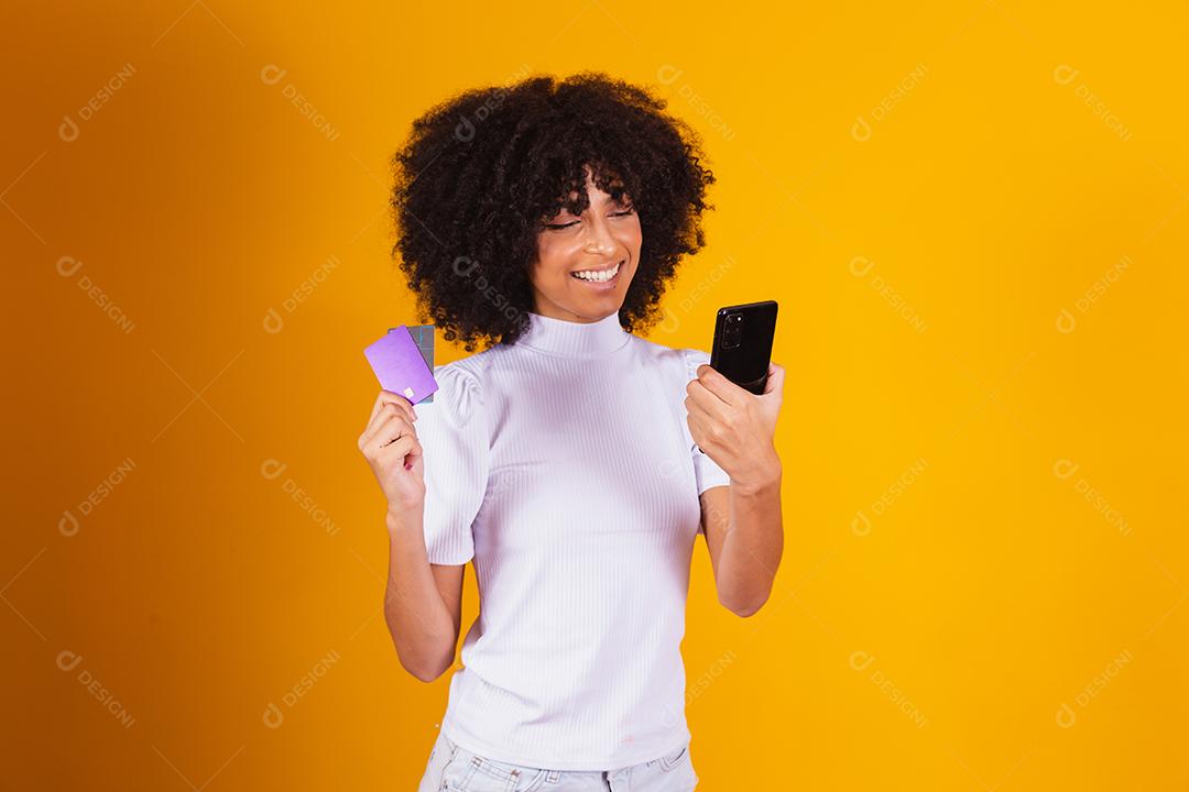 Foto de linda mulher de pele escura animada segurando cartão de crédito plástico mostrando novo modelo de telefone