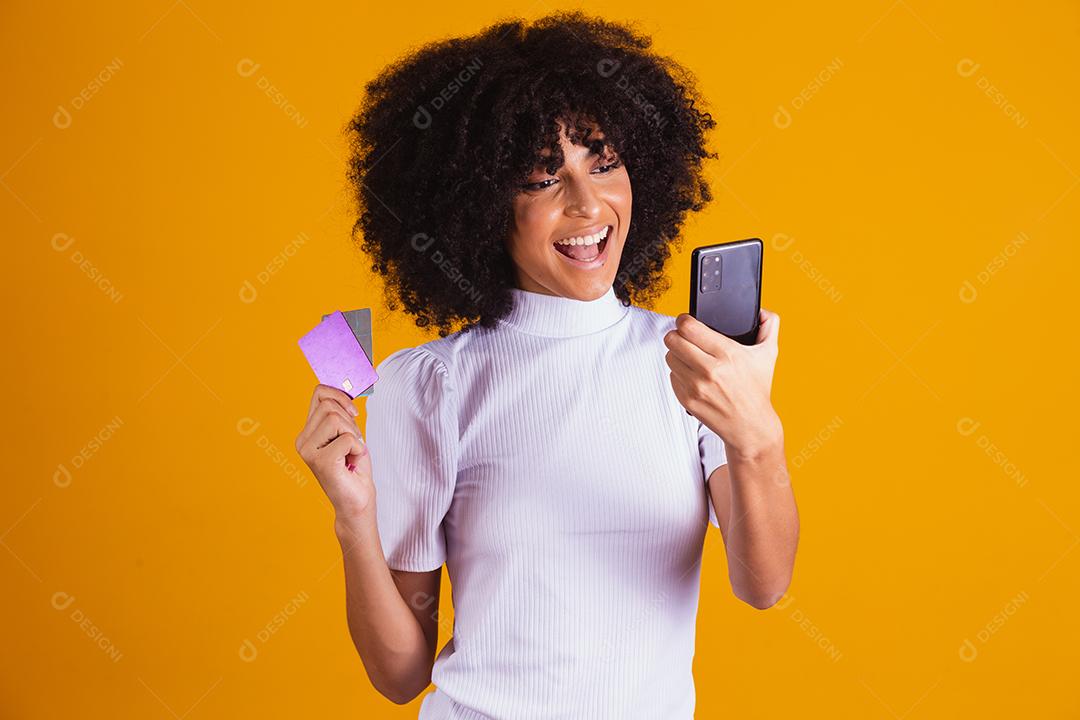 Foto de linda mulher de pele escura animada segurando cartão de crédito plástico mostrando novo modelo de telefone