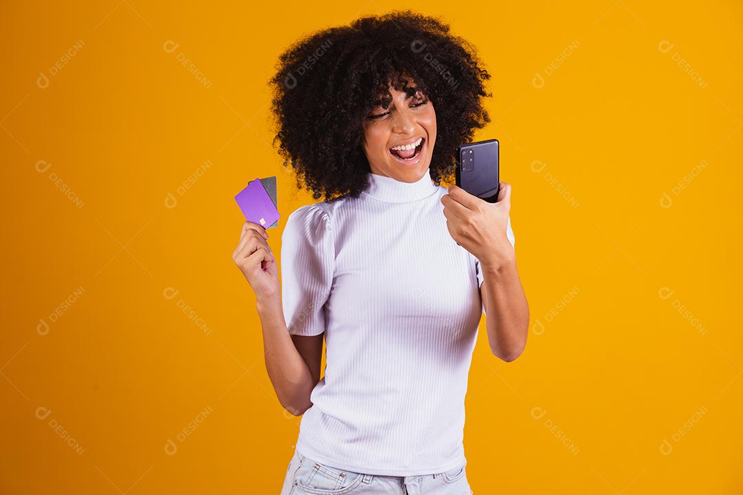 Foto de linda mulher de pele escura animada segurando cartão de crédito plástico mostrando novo modelo de telefone