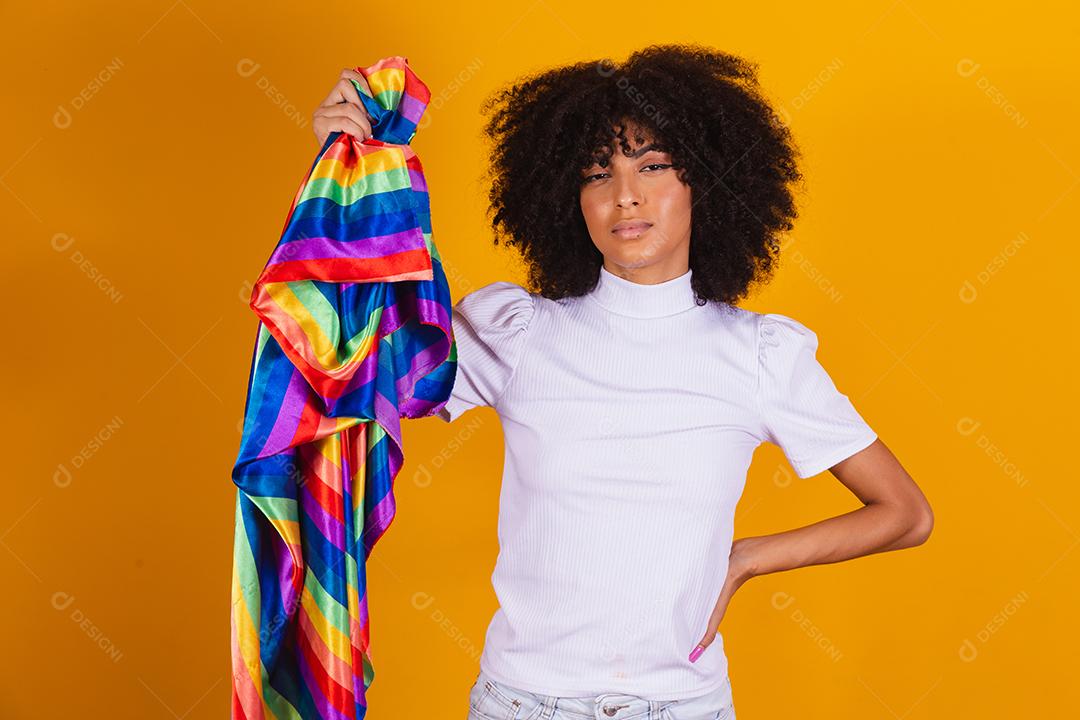 Mulher afro com uma bandeira lgbt nas mãos cobre metade do rosto com a outra mão. Mulher lesciba segurando uma bandeira lgbt