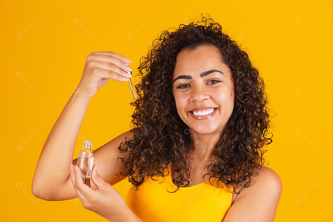 Tratamento reparador de pontas duplas. Mulher africana sorridente pulverizando óleo essencial em seu cabelo castanho encaracolado