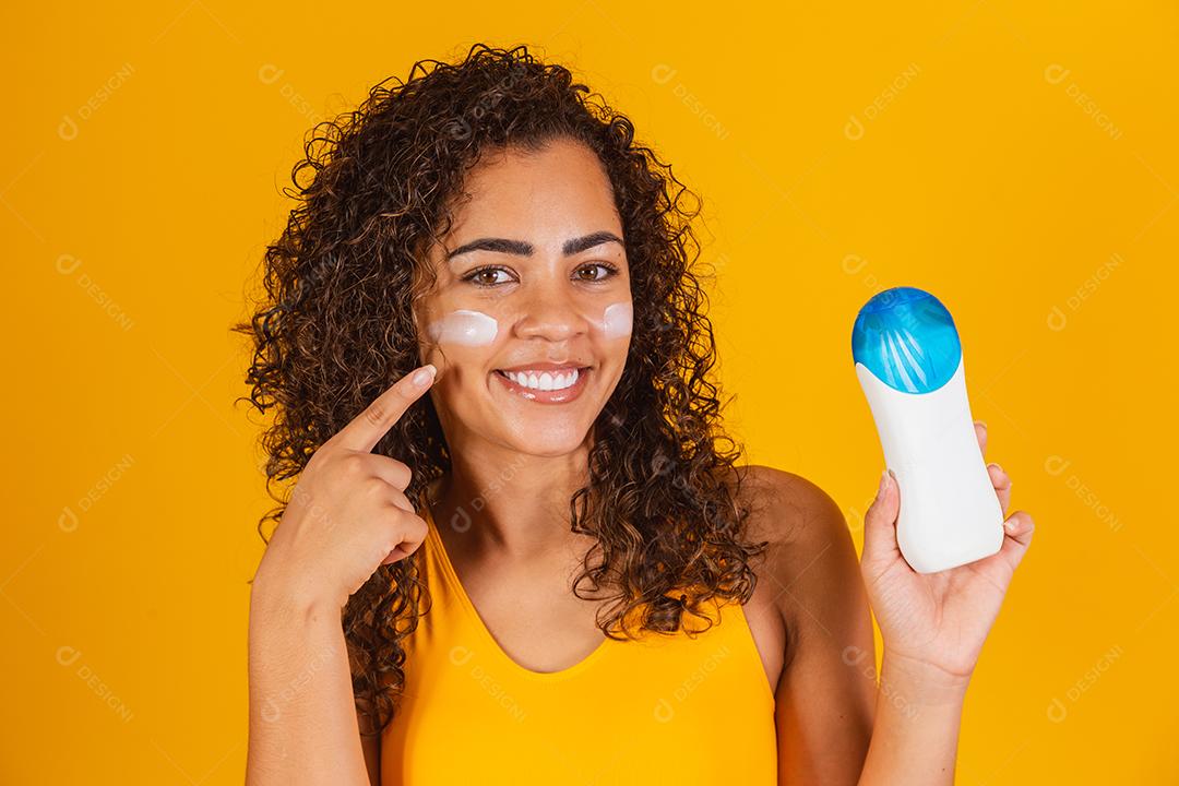 Jovem de maiô segurando um protetor solar. Jovem afro colocando protetor solar para proteger do sol