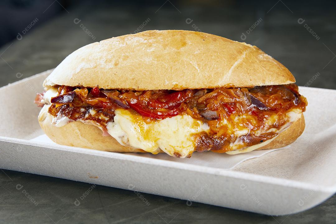 Sanduíche de carne, queijo derretido, cebola e pimenta em um prato branco sobre fundo de pedra.