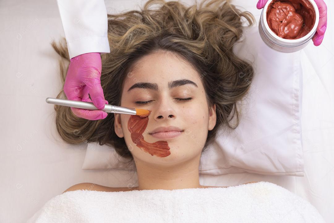 mulher está deitada no consultório do cosmetologista sobre o procedimento de hidratação do rosto. O cosmetologista aplica uma máscara hidratante no rosto do paciente.