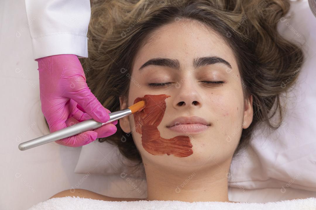 mulher está deitada no consultório do cosmetologista sobre o procedimento de hidratação do rosto. O cosmetologista aplica uma máscara hidratante no rosto do paciente.