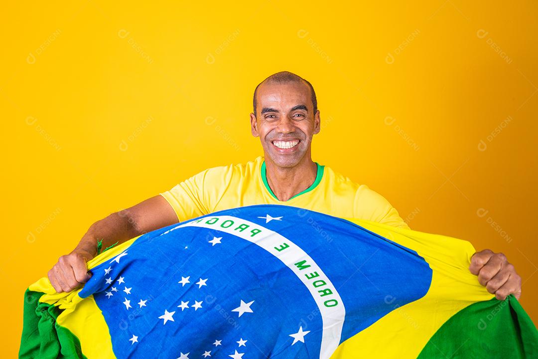 Jogador de futebol brasileiro comemorando em um fundo amarelo. Torcedor brasileiro de homem afro comemorando segurando a bandeira do brasil