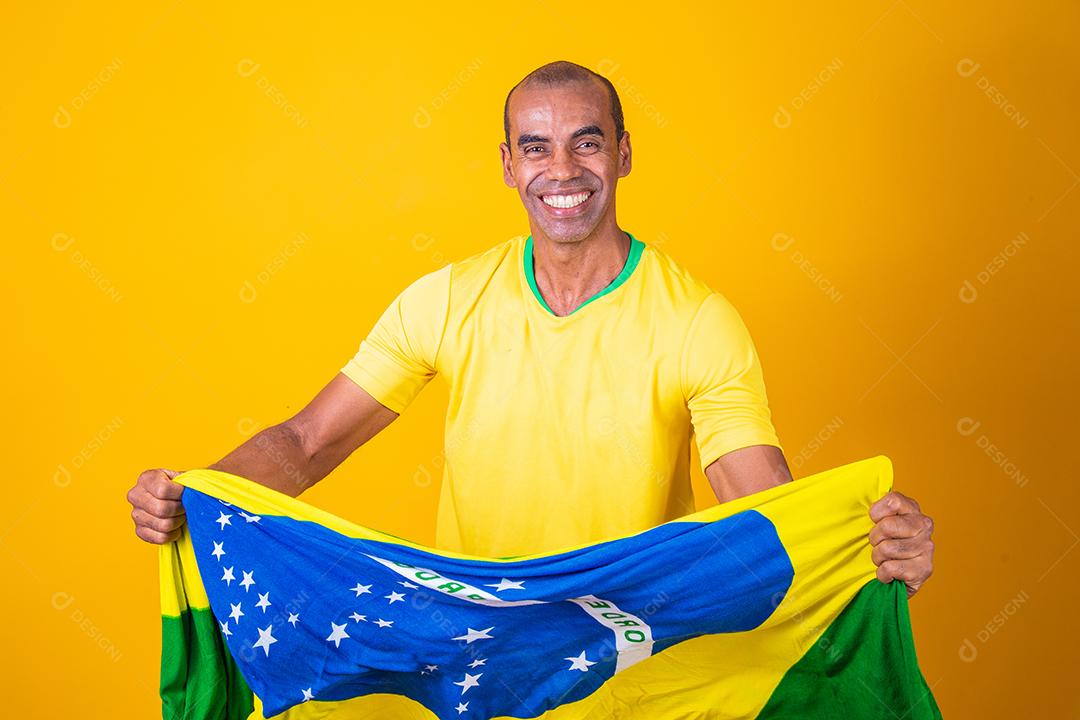 Jogador de futebol brasileiro comemorando em um fundo amarelo. Torcedor brasileiro de homem afro comemorando segurando a bandeira do brasil