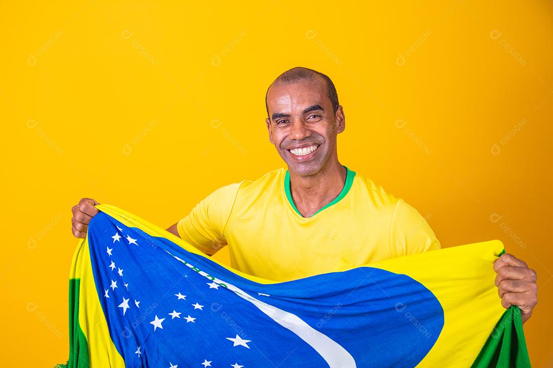 Jogador de futebol brasileiro comemorando em um fundo amarelo. Torcedor brasileiro de homem afro comemorando segurando a bandeira do brasil