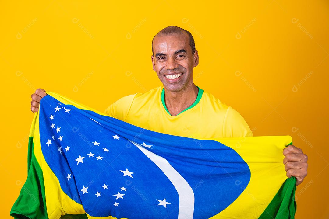 Jogador de futebol brasileiro comemorando em um fundo amarelo. Torcedor brasileiro de homem afro comemorando segurando a bandeira do brasil