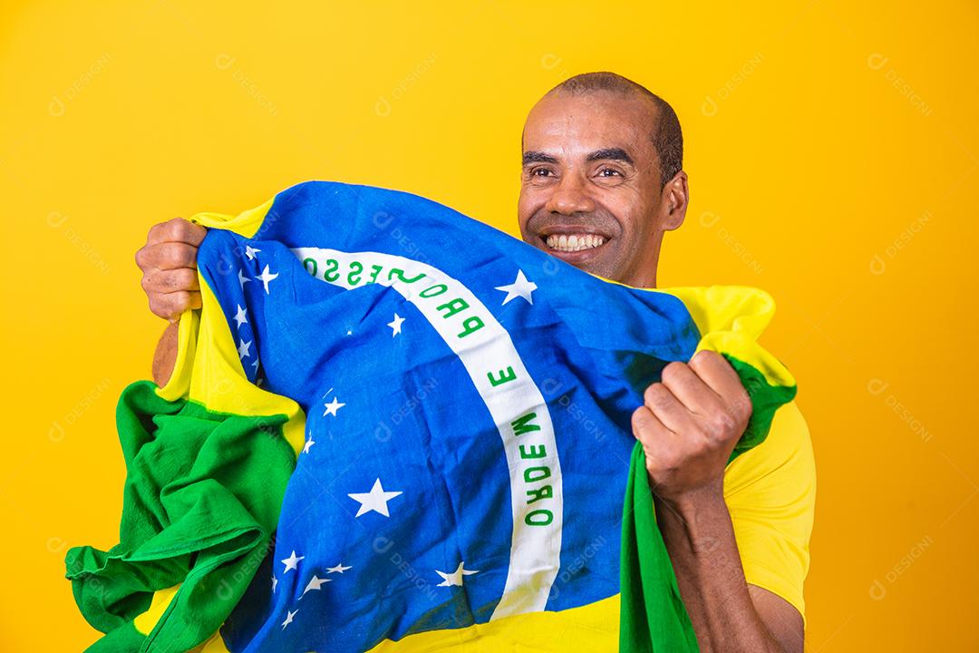 Jogador de futebol brasileiro comemorando em um fundo amarelo. Torcedor brasileiro de homem afro comemorando segurando a bandeira do brasil