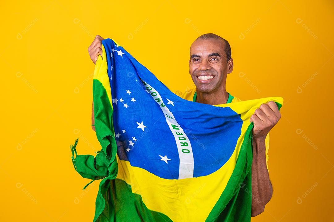 Jogador de futebol brasileiro comemorando em um fundo amarelo. Torcedor brasileiro de homem afro comemorando segurando a bandeira do brasil