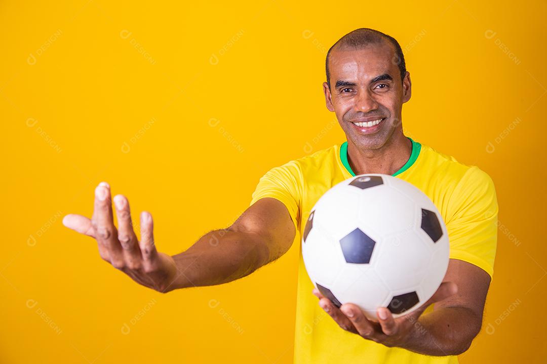Homem fã brasileiro segurando uma bola de futebol em fundo amarelo
