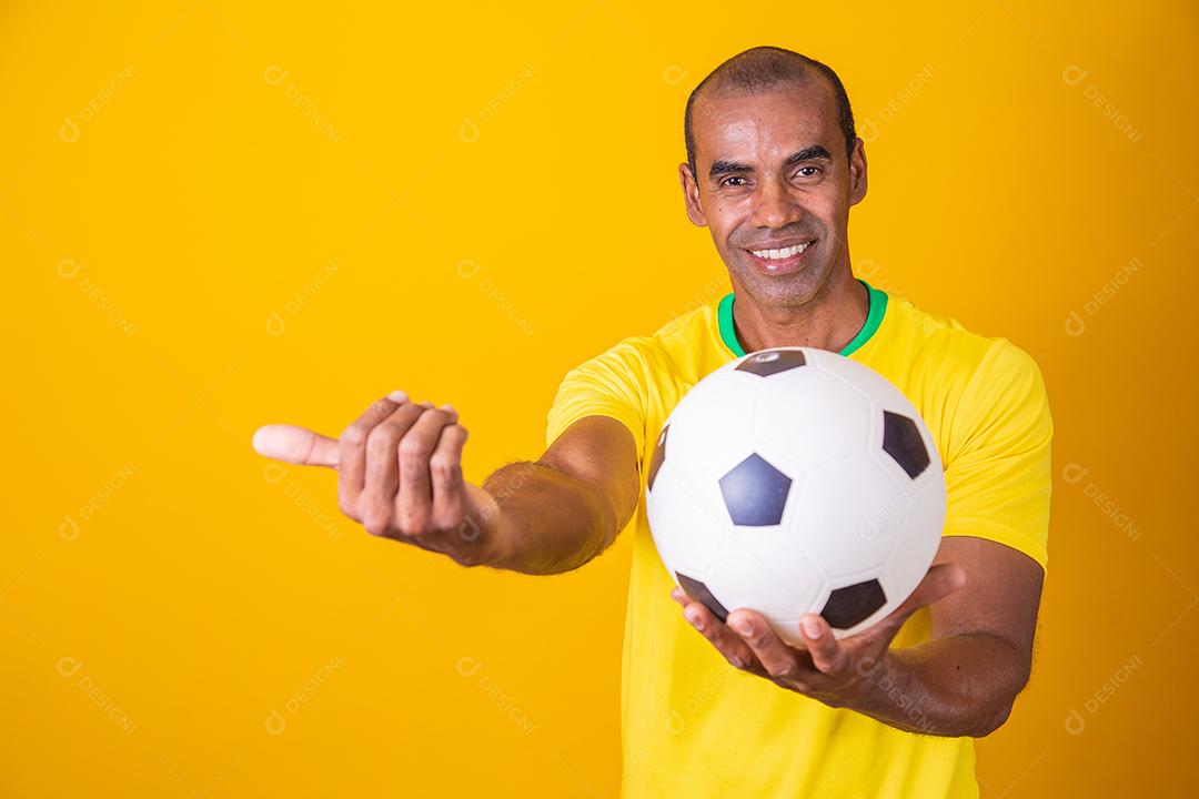 Homem fã brasileiro segurando uma bola de futebol em fundo amarelo