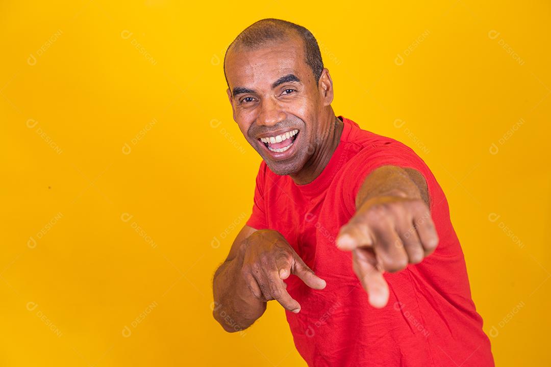 Homem negro positivo aponta o dedo indicador para a câmera, sorri alegremente e escolhe alguém isolado em fundo amarelo