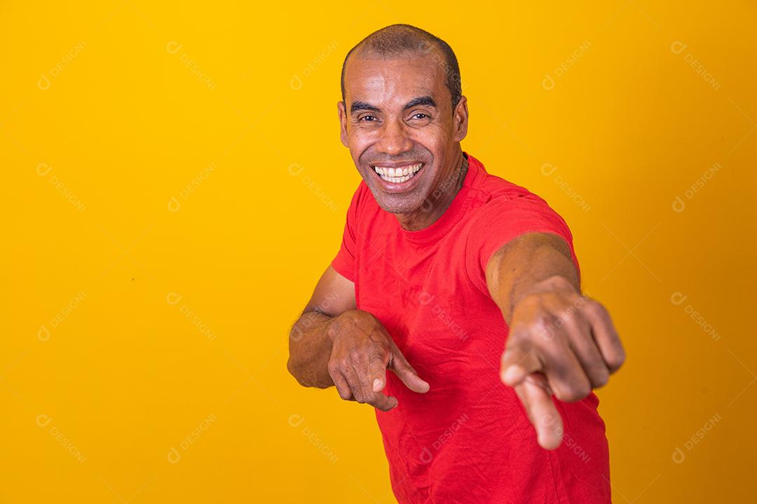 Homem negro positivo aponta o dedo indicador para a câmera, sorri alegremente e escolhe alguém isolado em fundo amarelo