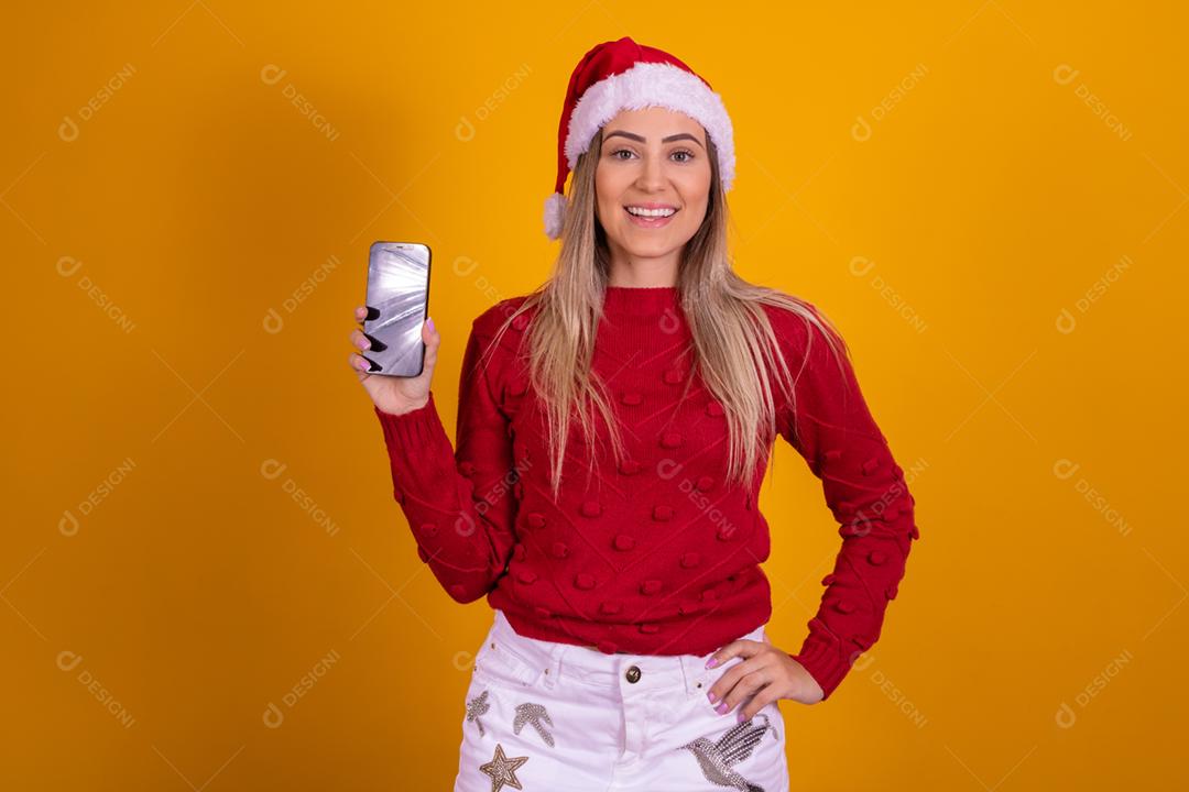 Mulher loira com um chapeu de papai noel segurando um celular em fundo amarelo