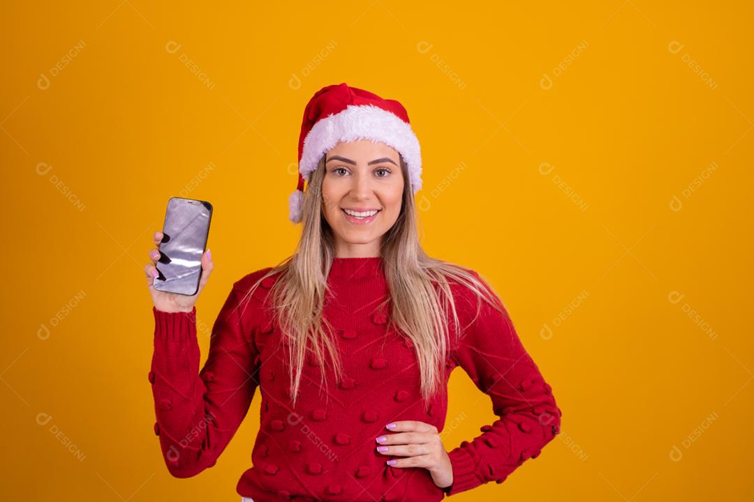 Mulher loira com um chapeu de papai noel segurando um celular em fundo amarelo