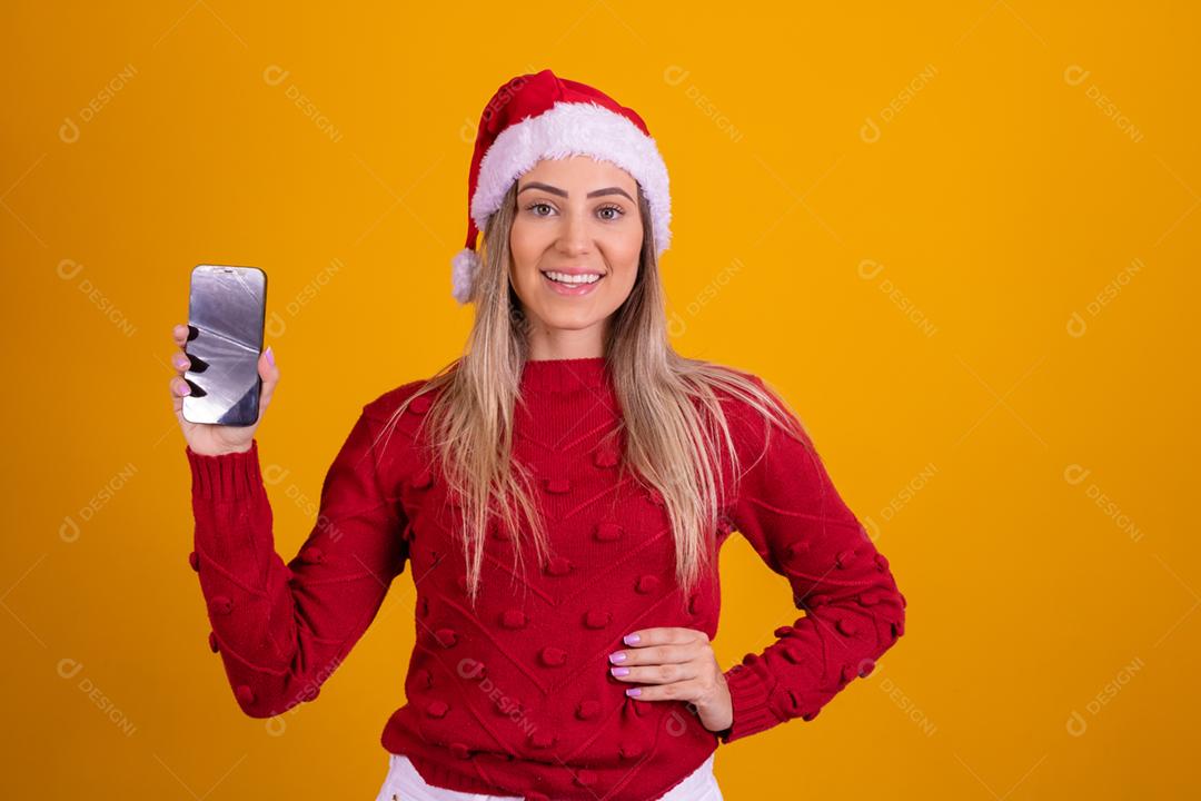 Mulher loira com um chapeu de papai noel segurando um celular em fundo amarelo