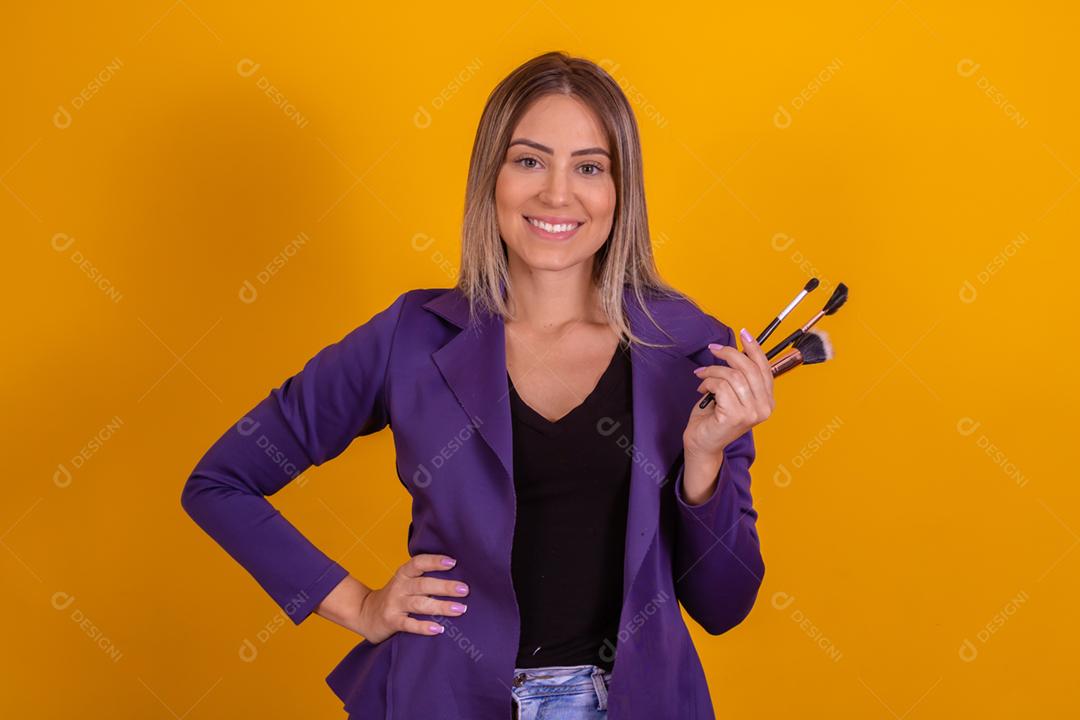 Mulher loira com um blazer roxo segurando pinceis de maquiagem