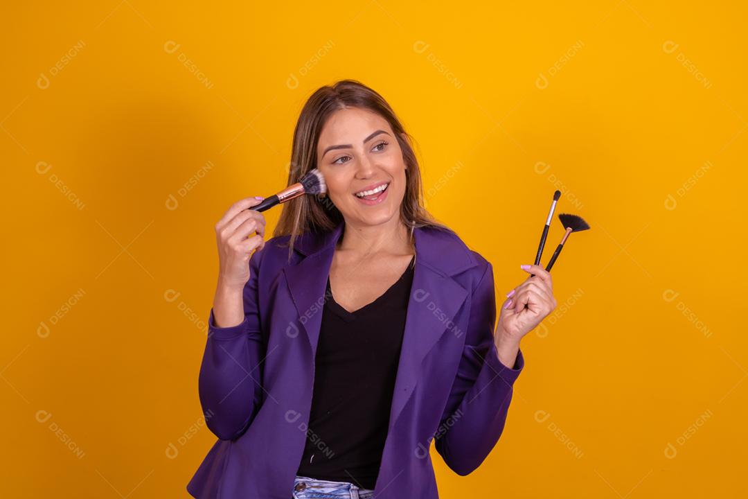 Mulher loira com um blazer roxo segurando pinceis de maquiagem