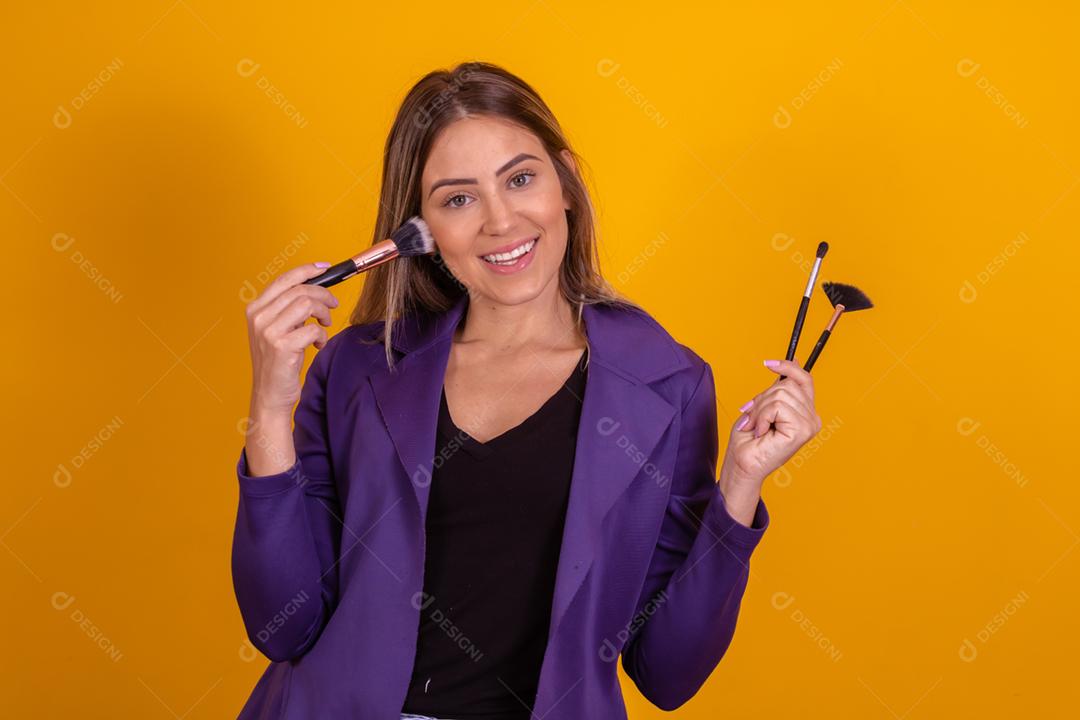 Mulher loira com um blazer roxo segurando pinceis de maquiagem