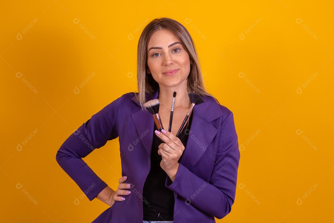 Mulher loira com um blazer roxo segurando pinceis de maquiagem